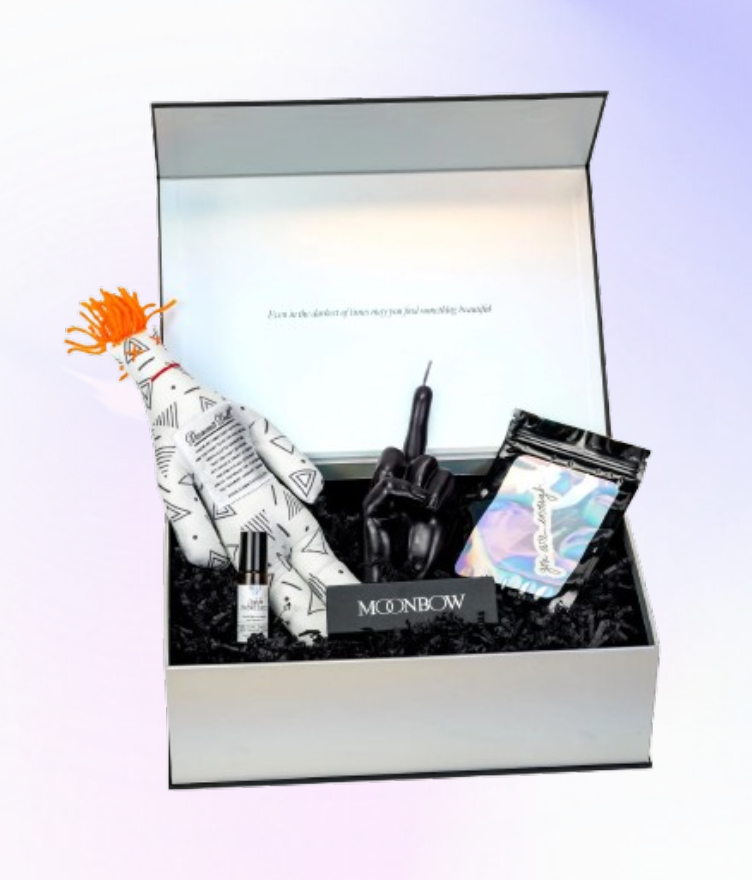 A Moonbow gift box with stress relief items, including a plush doll, black candle, essential oil roller, and holographic packaging.